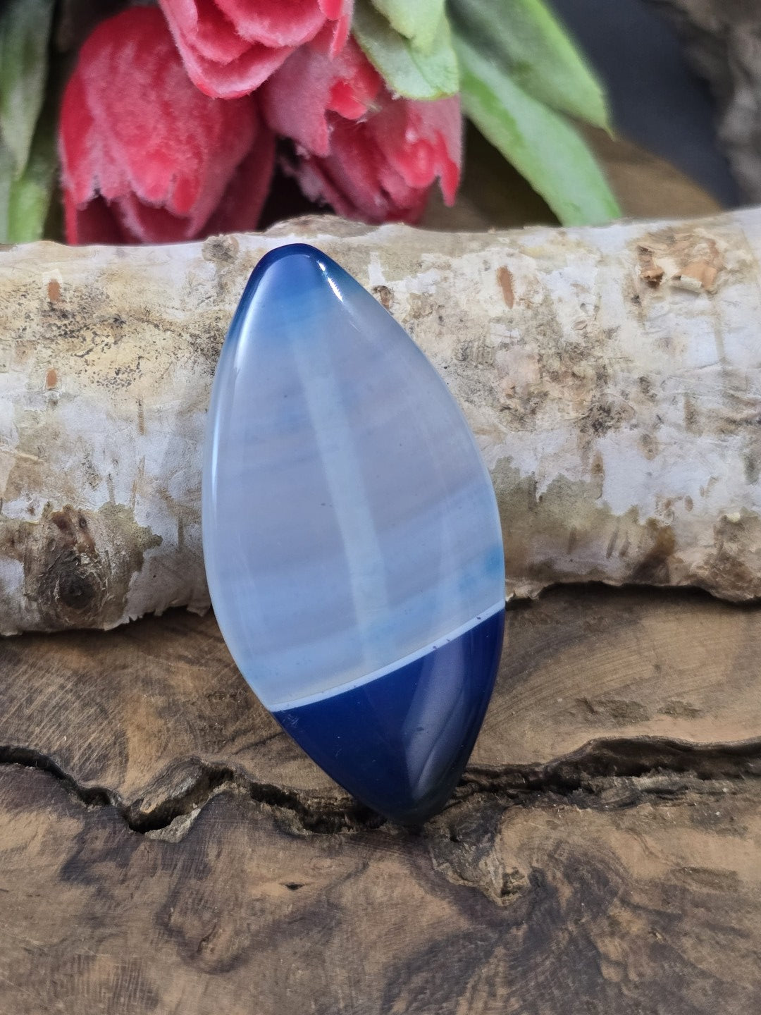 Blaue Achat-Centerperle (39 mm) in Navette-Form mit zweifarbiger Bänderung auf rustikalem Untergrund vor einem Birkenstamm.