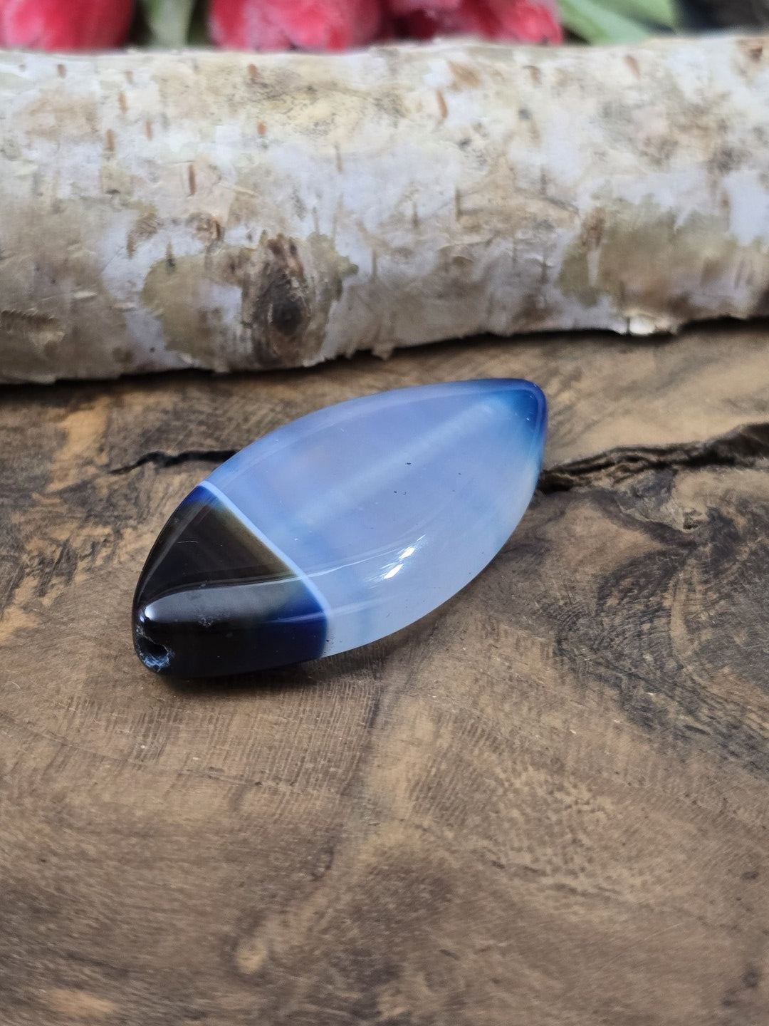 Liegende blaue Achatperle in Navette-Form auf Holz zur Verdeutlichung der Wölbung und der seitlichen Fädelbohrung.