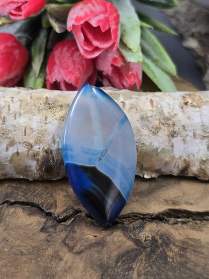 Blaue Achat-Centerperle (39 mm) in Navette-Form mit feiner Edelstein-Maserung und dunklem Farbverlauf vor einem Birkenstamm.