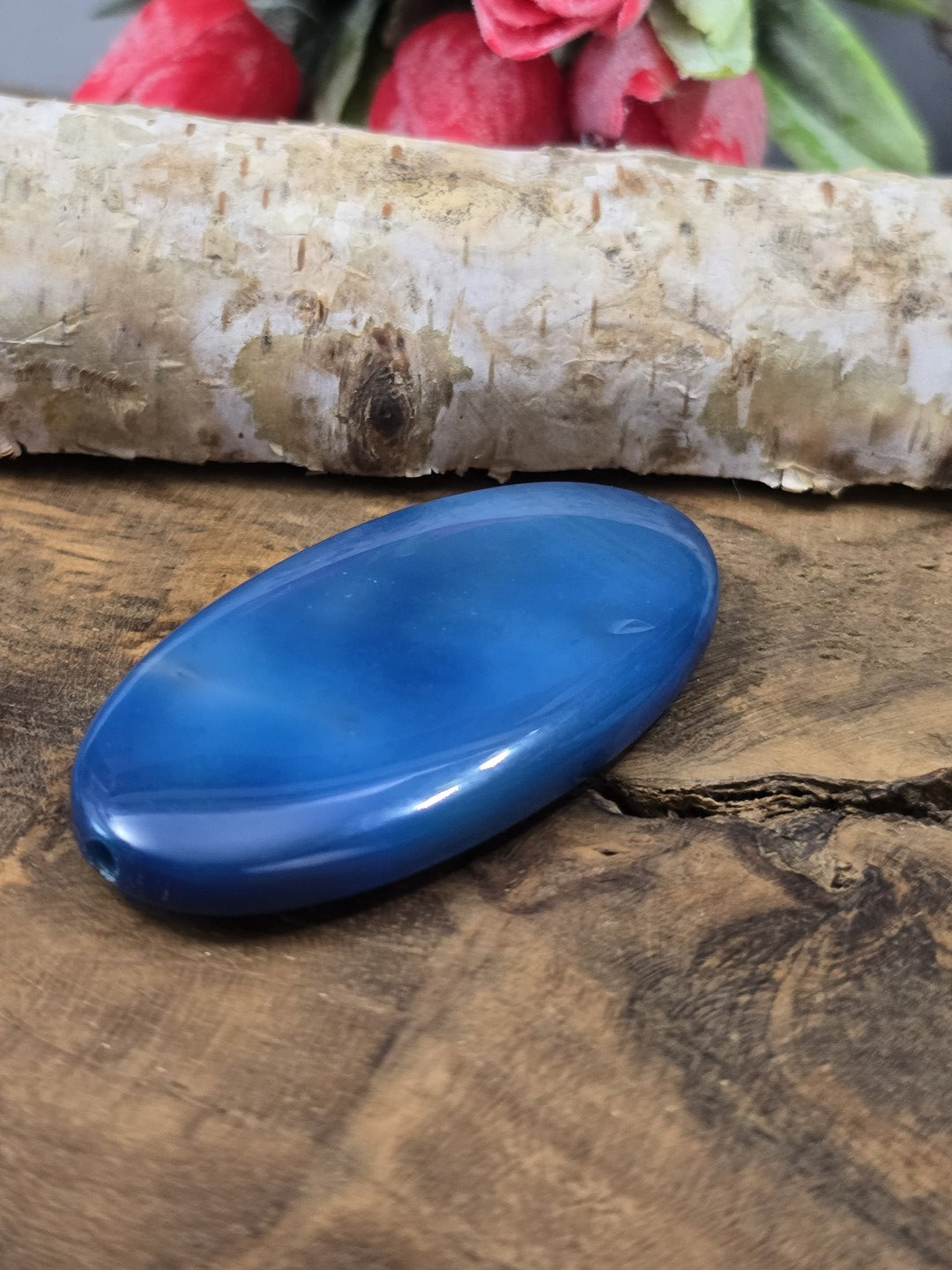 Liegende blaue Achat-Centerperle (50 mm) auf Holz zur Verdeutlichung der Wölbung und der Fädelbohrung.