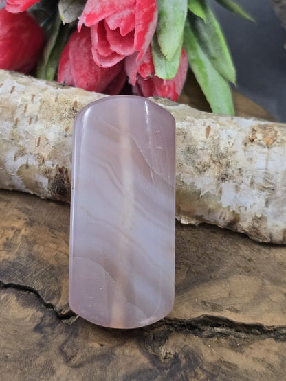 Zartrosa Achat-Centerperle (50 mm) in Pillow-Rechteckform mit feiner Bänderung vor Birkenstamm und roten Blüten.
