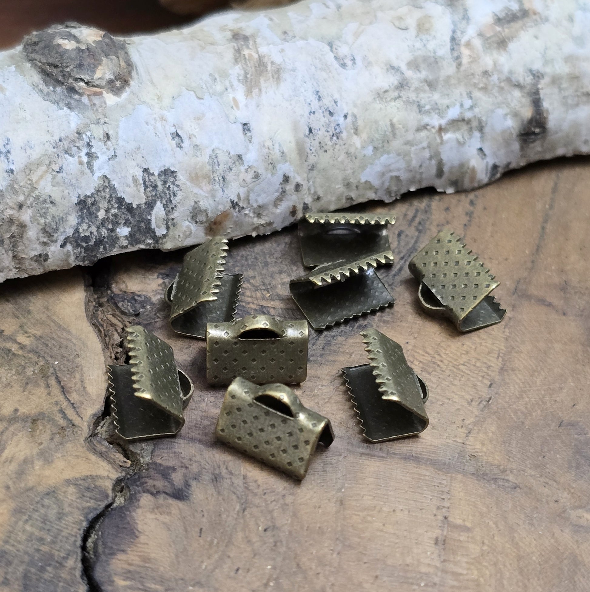 Das Bild zeigt antik bronzefarbige Bandklemmen mit Zähnen, 10 mm breit – ideal zum Befestigen von Verschlüssen an Stoffbändern. Einfach mit einer Zange auf das Bandende klemmen.