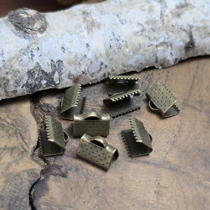 Das Bild zeigt antik bronzefarbige Bandklemmen mit Zähnen, 10 mm breit – ideal zum Befestigen von Verschlüssen an Stoffbändern. Einfach mit einer Zange auf das Bandende klemmen.