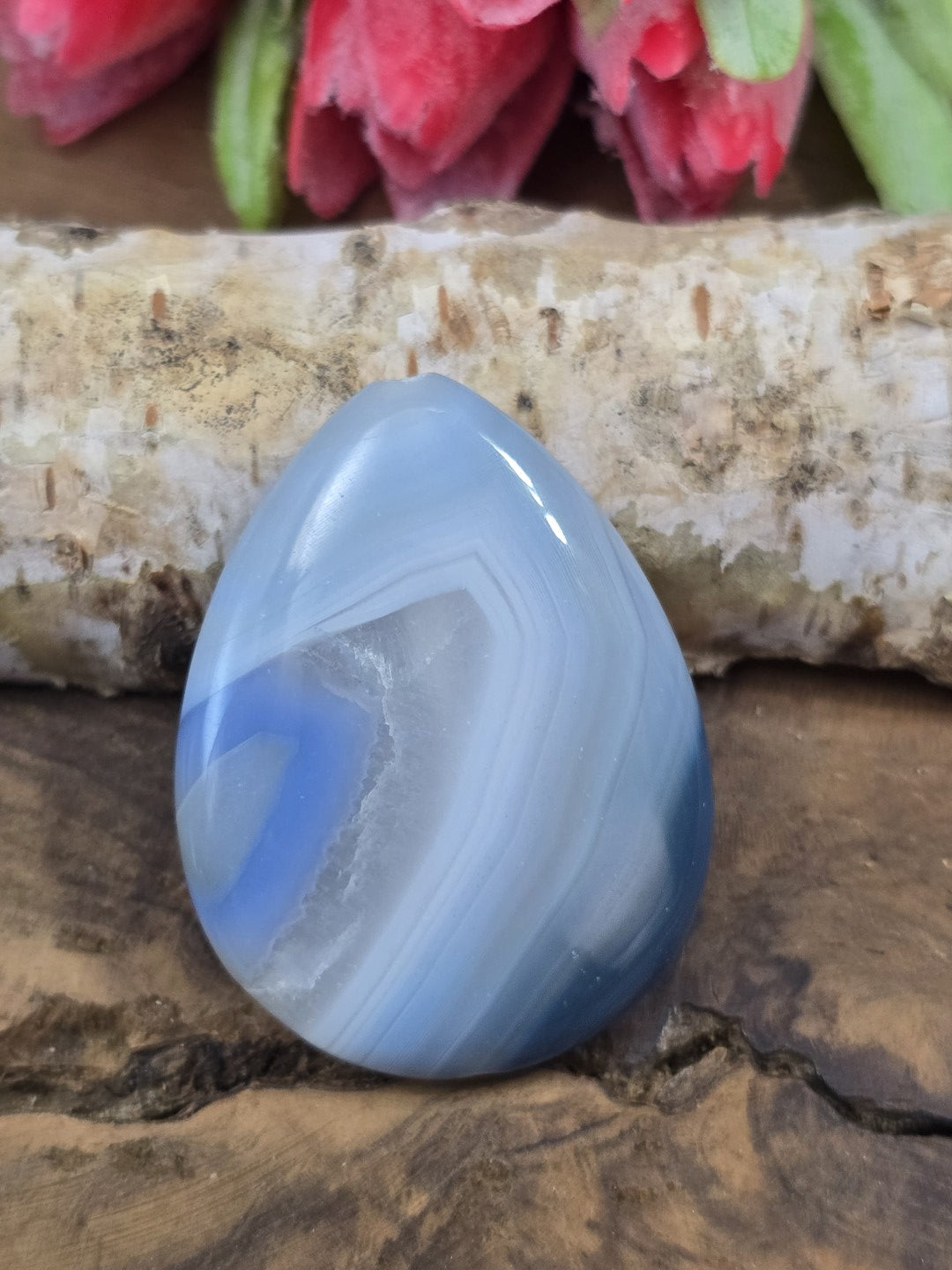 Vorderseite eines blauen Tropfen-Achat-Edelsteins mit natürlicher Bänderung auf Rustikal-Holzgrund vor Birkenholz.