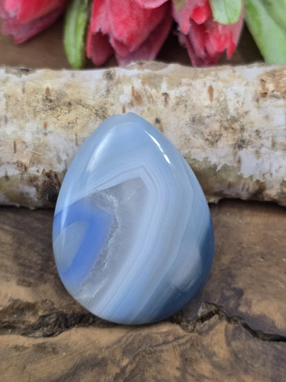 Vorderseite eines blauen Tropfen-Achat-Edelsteins mit natürlicher Bänderung auf Rustikal-Holzgrund vor Birkenholz.