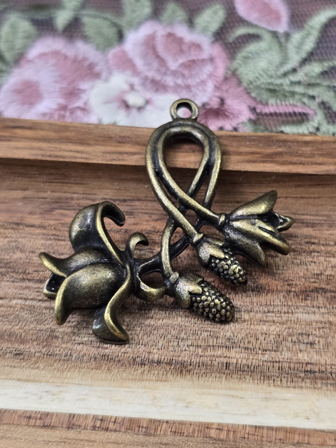 Detaillierter Blumen-Anhänger in Antikbronze mit filigranen Blüten und Öse flach liegend auf Holz.