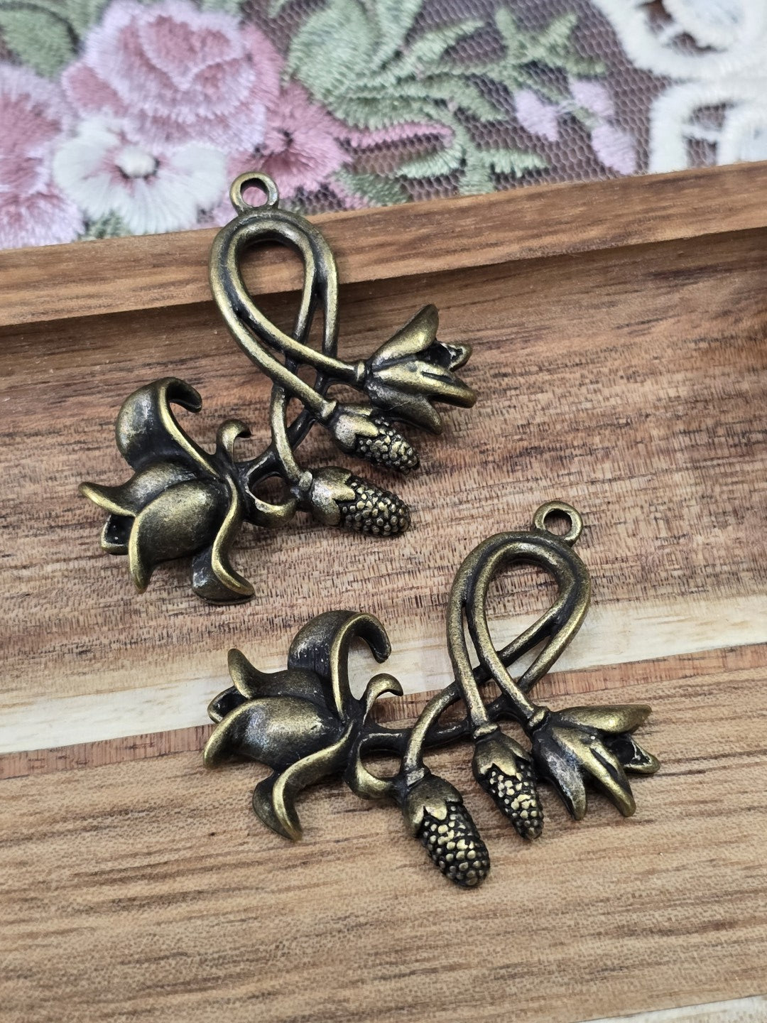 Zwei florale Schmuckanhänger mit Blüten-Motiv in Antikbronze nebeneinander auf Holzgrund.
