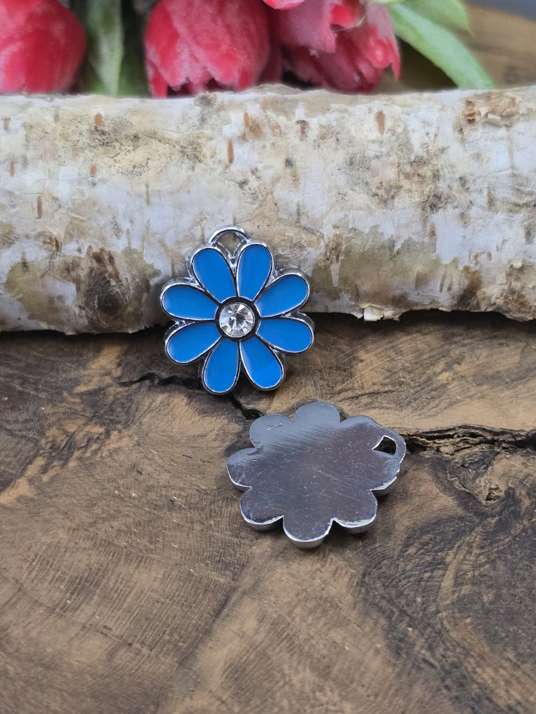 Ein blauer Blumenanhänger aus Edelstahl und Emaille mit Strassstein neben einem zweiten Anhänger, der die flache, silberfarbene Rückseite zeigt.