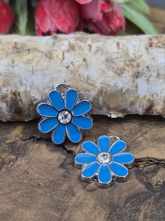 Zwei hellblaue Blumen-Schmuckanhänger aus Edelstahl mit funkelndem Strass-Zentrum dekorativ auf rustikalem Holz vor Birkenrinde.
