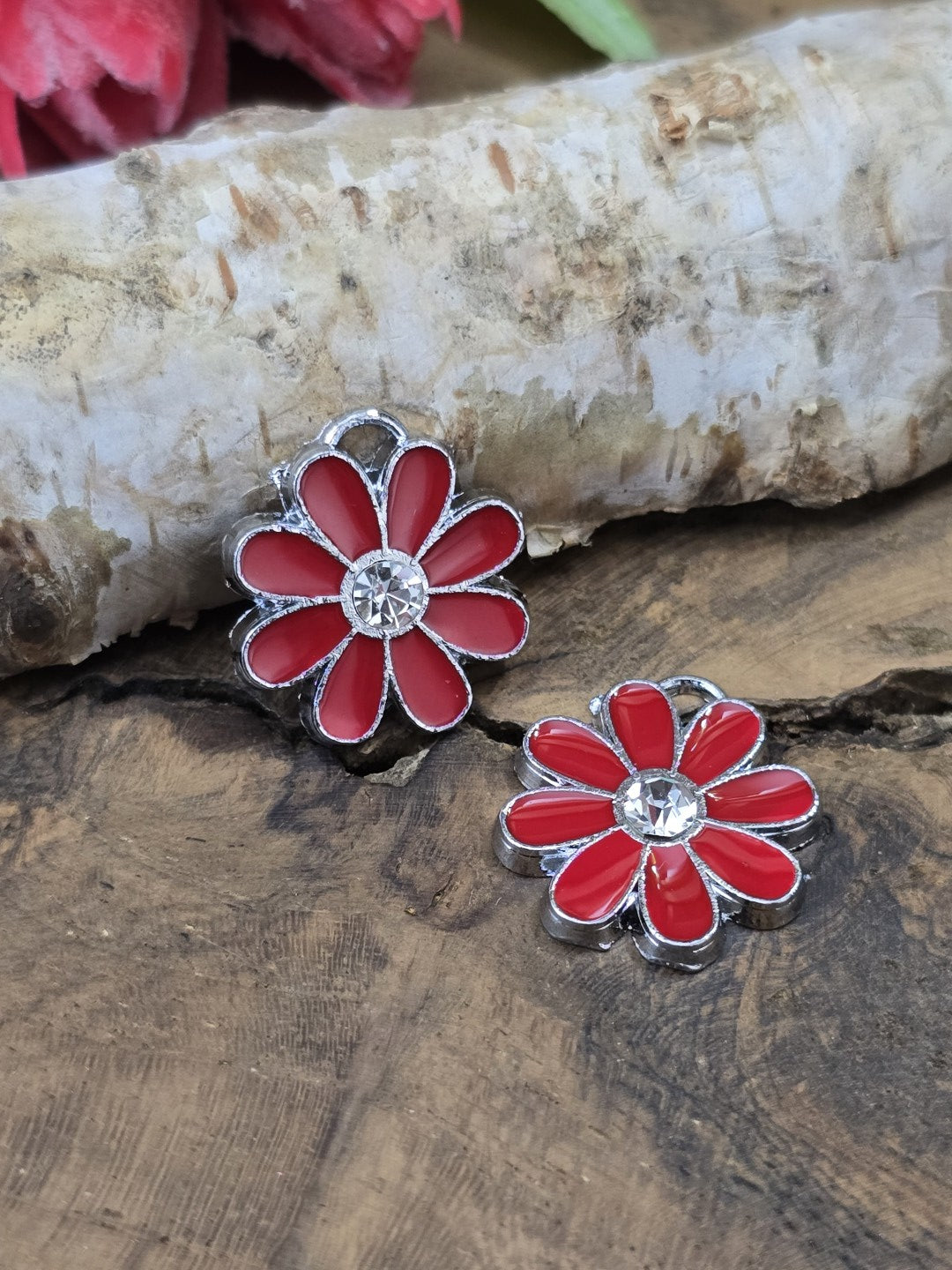 Zwei rote Blumenanhänger aus Edelstahl und Emaille mit glitzerndem Strassstein im Zentrum auf einer Holzoberfläche.