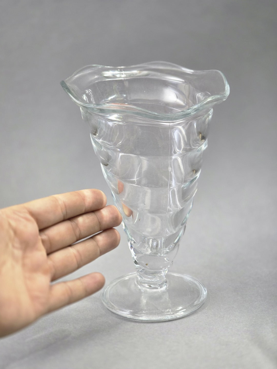 Eine Hand neben dem transparenten Eisbecher zeigt die Größe, während Schale, runder Fuß, Spiraldekor und gewellter Rand gut erkennbar sind.