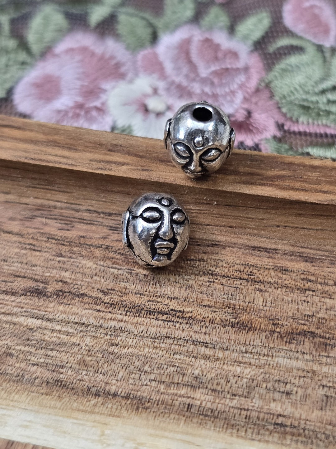Zwei Buddha-Kopf Perlen in Antiksilber flach liegend auf Holzgrund.