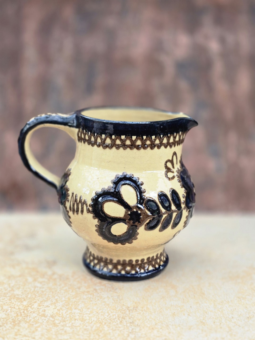 Vintage Keramikkrug im Bunzlau Stil mit cremefarbener Glasur, dunklem floralem Dekor, bauchiger Form, kleinem Ausguss und geschwungenem Henkel. Die Kanne zeigt einen hellen, dichten Scherben und wirkt wie handwerkliche Volkskeramik aus der ersten Hälfte des 20. Jahrhunderts. Guter Vintagezustand mit kleinen Chips am Ausguss und einem kleinen Chip am Boden.