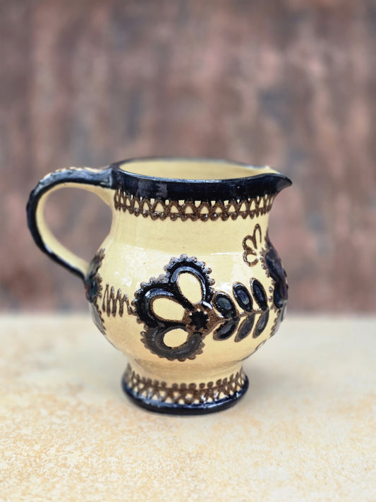 Vintage Keramikkrug im Bunzlau Stil mit cremefarbener Glasur, dunklem floralem Dekor, bauchiger Form, kleinem Ausguss und geschwungenem Henkel. Die Kanne zeigt einen hellen, dichten Scherben und wirkt wie handwerkliche Volkskeramik aus der ersten Hälfte des 20. Jahrhunderts. Guter Vintagezustand mit kleinen Chips am Ausguss und einem kleinen Chip am Boden.