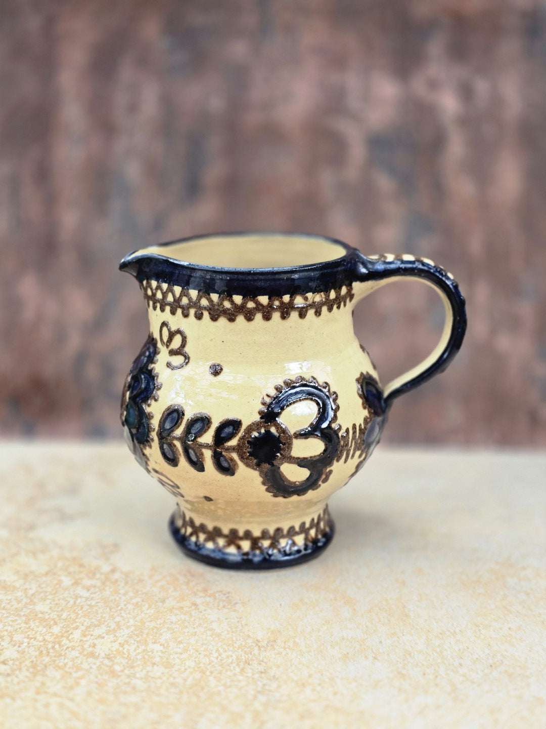 Vintage Keramikkanne mit bauchigem Korpus, kurzem Ausguss, geschwungenem Henkel und dunklem floralem Ornament auf cremefarbener Glasur.