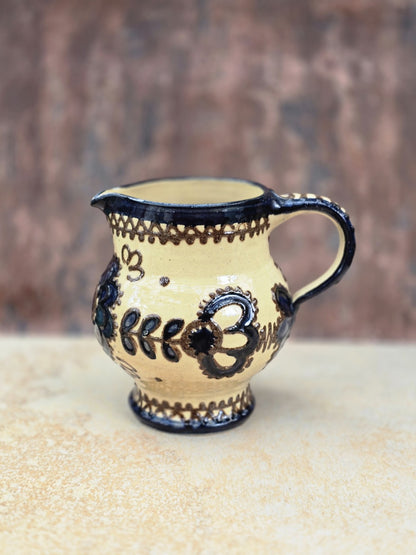 Vintage Keramikkanne mit bauchigem Korpus, kurzem Ausguss, geschwungenem Henkel und dunklem floralem Ornament auf cremefarbener Glasur.