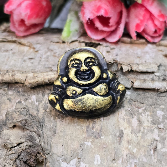 Das Bild zeigt einen handgefertigten Cabochon in Form eines lachenden Buddhas aus Kunstharz. Der Buddha hat eine schwarze Oberfläche mit bronzefarbenen Details und ein kleines Loch am oberen Rand. Die Rückseite ist flach, ideal für die Verwendung als Anhänger oder in Bastelprojekten.