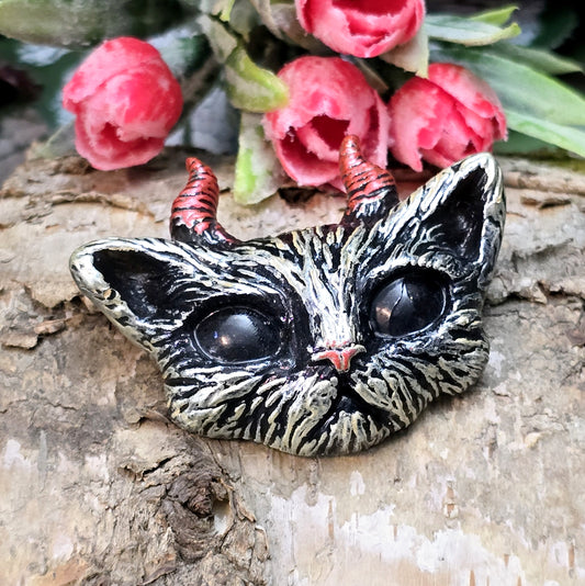 Auf dem Bild ist ein handgefertigter Katzenkopf mit Hörnern aus Kunstharz zu sehen. Der Kopf ist mit handbemalten Details verziert und hat eine flache Rückseite, die ideal für Schmuckherstellung oder Bastelprojekte geeignet ist.