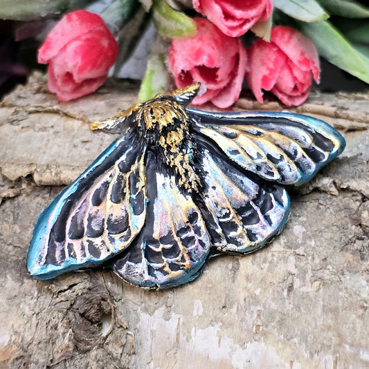 Auf dem Bild ist ein handgefertigter Cabochon in Form einer Deathmoth-Motte zu sehen. Das detailreiche Kunstharzstück zeigt die ausgebreiteten Flügel der mystischen Motte, verziert mit feinen Mustern und Schattierungen. Die Farbgebung betont die Struktur, wodurch das Motiv lebendig wirkt. Die Rückseite ist flach, sodass es sich gut für Schmuck oder Bastelarbeiten eignet. Die Motte liegt auf einem dunklen Untergrund, was ihren geheimnisvollen Charakter unterstreicht.