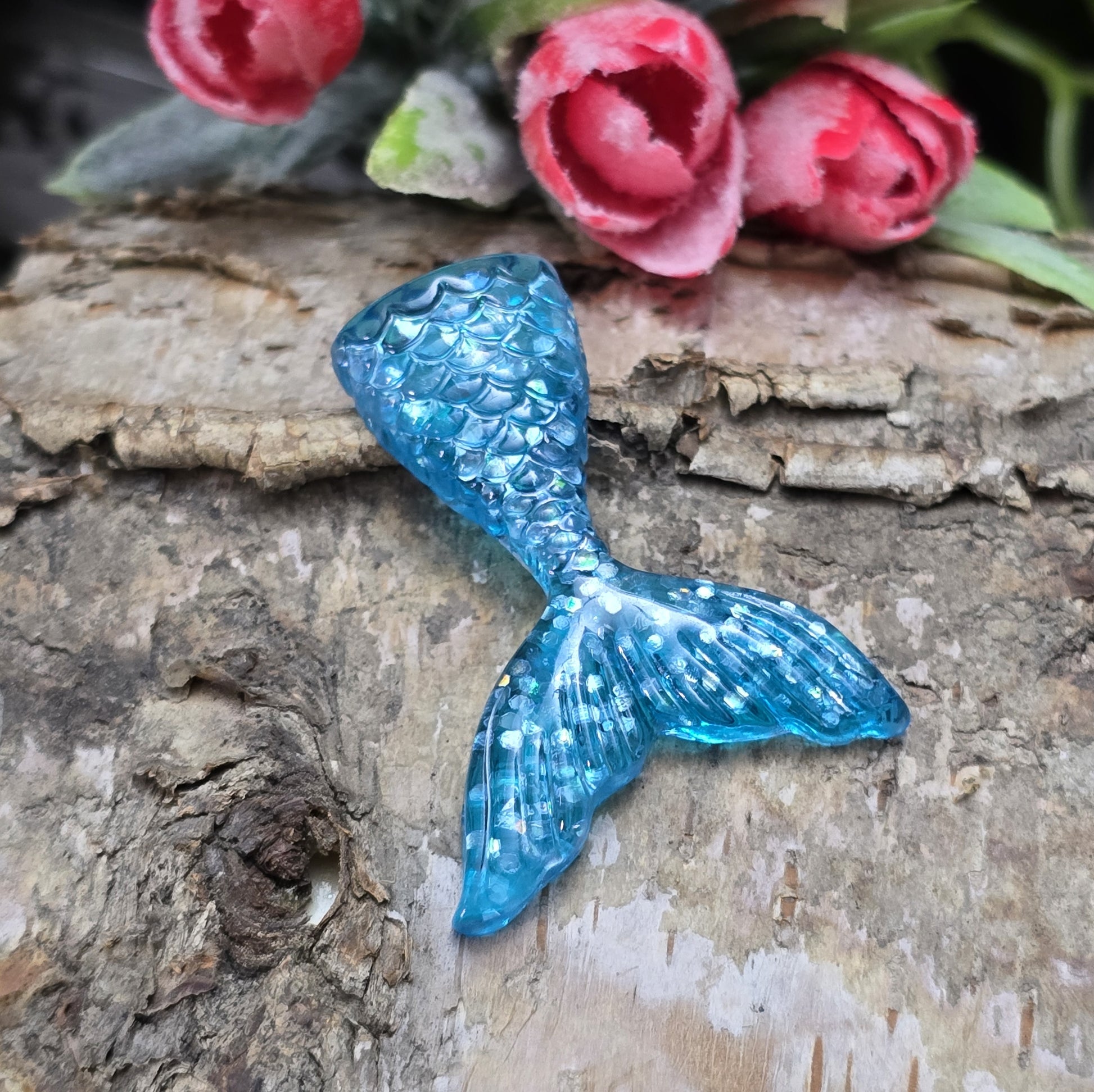 Das Bild zeigt einen handgefertigten Cabochon in Form einer Meerjungfrauenschwanzflosse. Der Anhänger besteht aus Kunstharz und leuchtet in einem schimmernden Aqua Blau mit feinem Glitter. Die geschwungene Form und die detailreiche Struktur verleihen ihm eine elegante, maritime Optik.