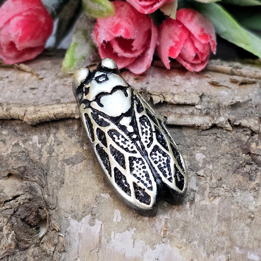 Das Bild zeigt einen handgefertigten Zikaden-Cabochon aus Kunstharz in schwarzer und rosé-silberner Farbgebung. Die Zikade hat eine flache Rückseite mit einem kleinen Loch zwischen den Augen, ideal für die Anbringung einer Anhängeröse.