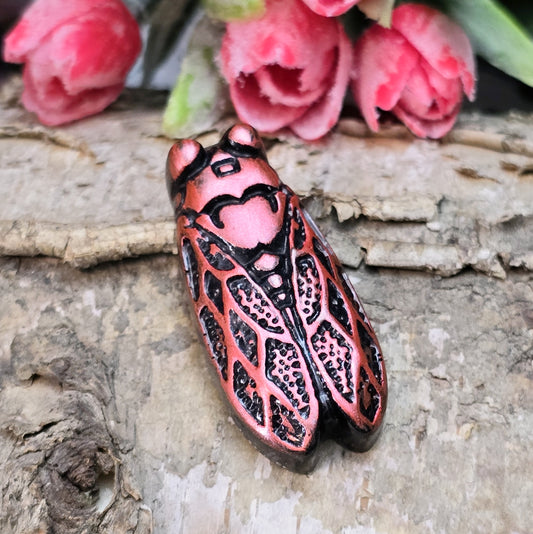Das Bild zeigt einen handgefertigten Zikaden-Cabochon aus Kunstharz in einer eleganten Karminrot-Optik. Die Zikade hat eine flache Rückseite mit einem kleinen Loch zwischen den Augen, das sich perfekt für die Anbringung einer Anhängeröse eignet.