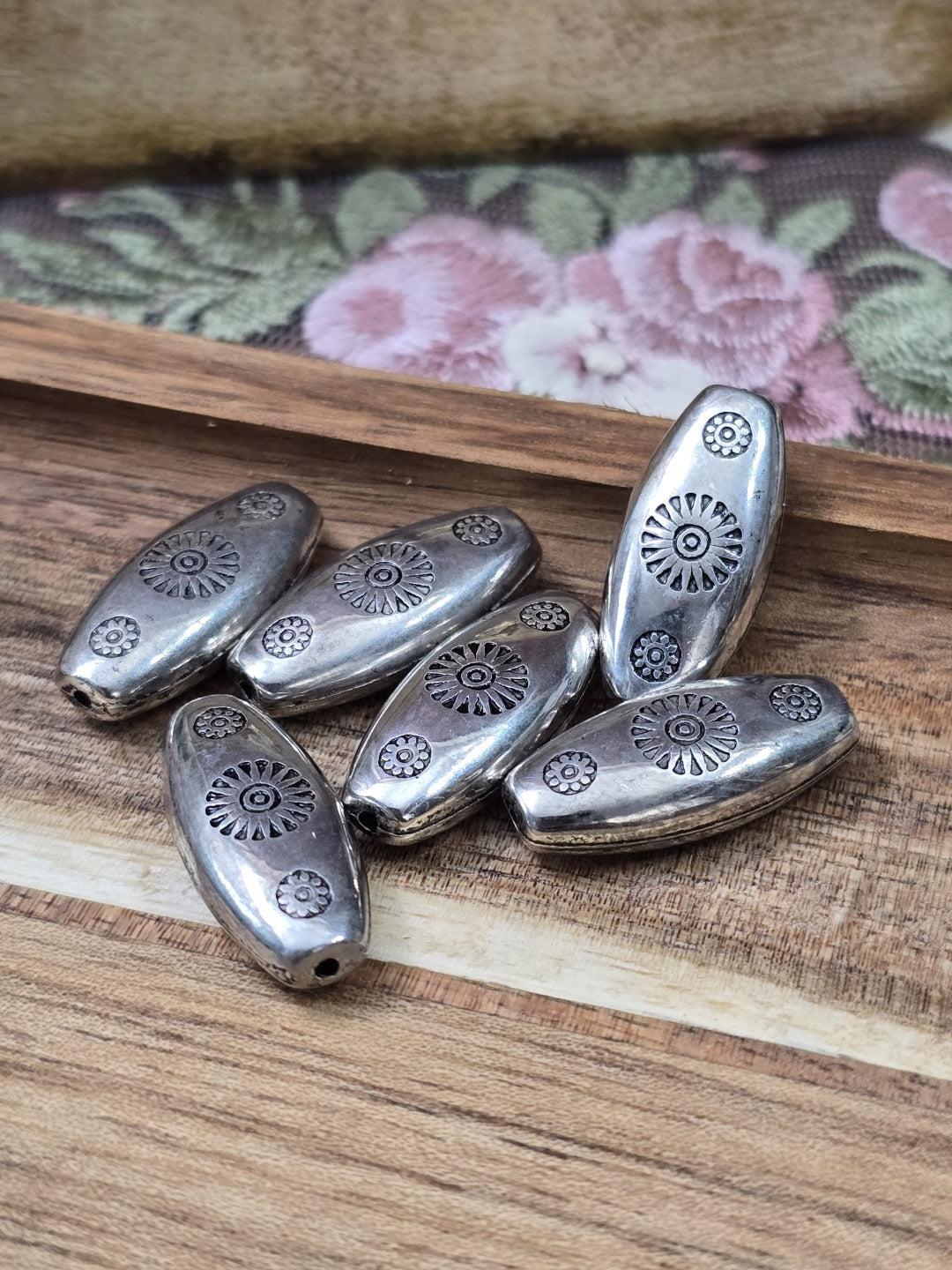 Oval langgezogene CCB-Perlen in Silber mit eingeprägtem Sonnen-Muster auf Holzuntergrund für DIY-Schmuck.