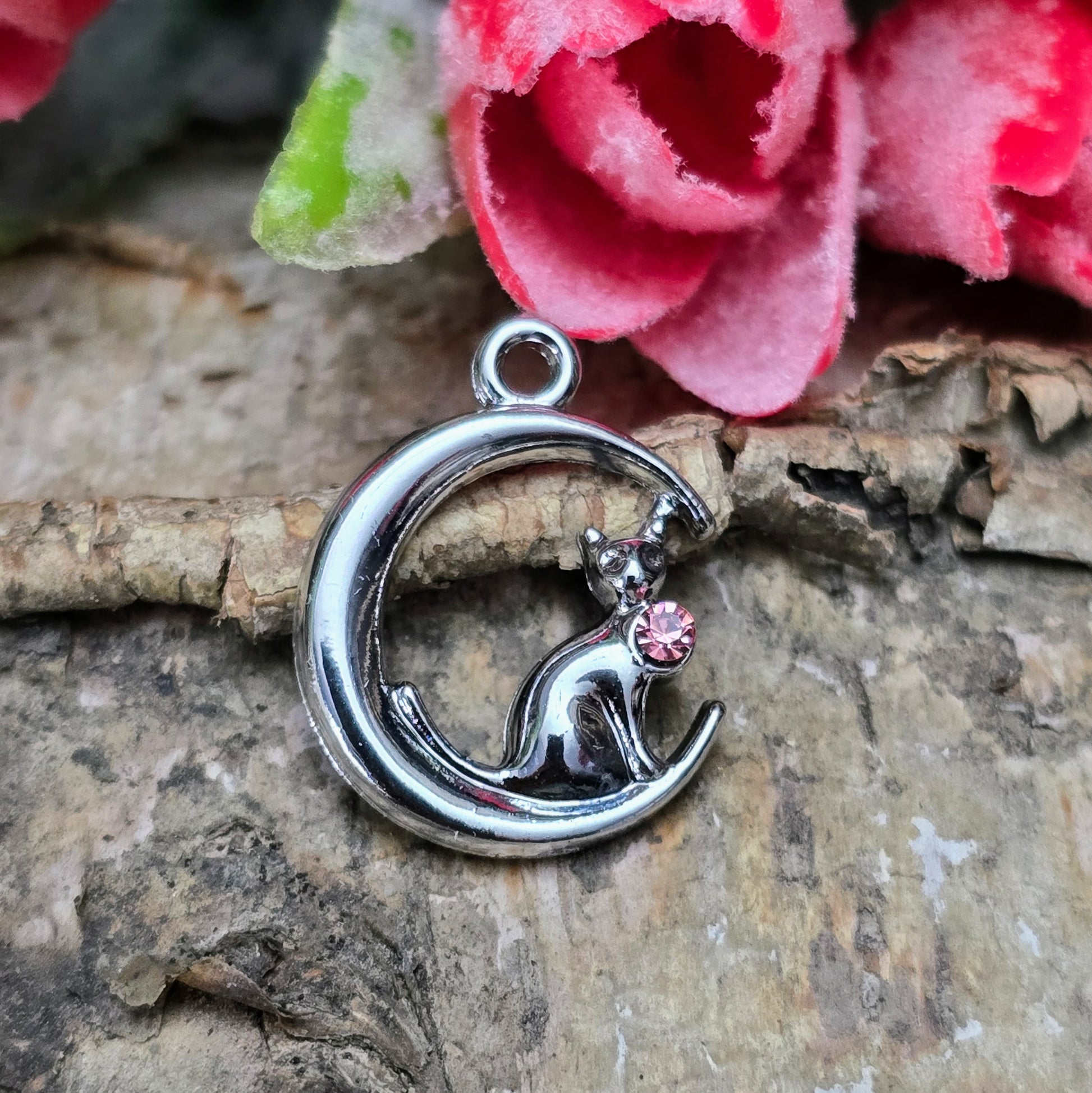 Das Bild zeigt einen Anhänger in Silberfarbe, der eine Katze im Mond darstellt. Die Katze ist elegant im Mond sitzend, begleitet von einem funkelnden rosé Strass-Stein. Der Anhänger ist ca. 20mm groß und eignet sich perfekt für die Schmuckherstellung oder als kreatives Accessoire.
