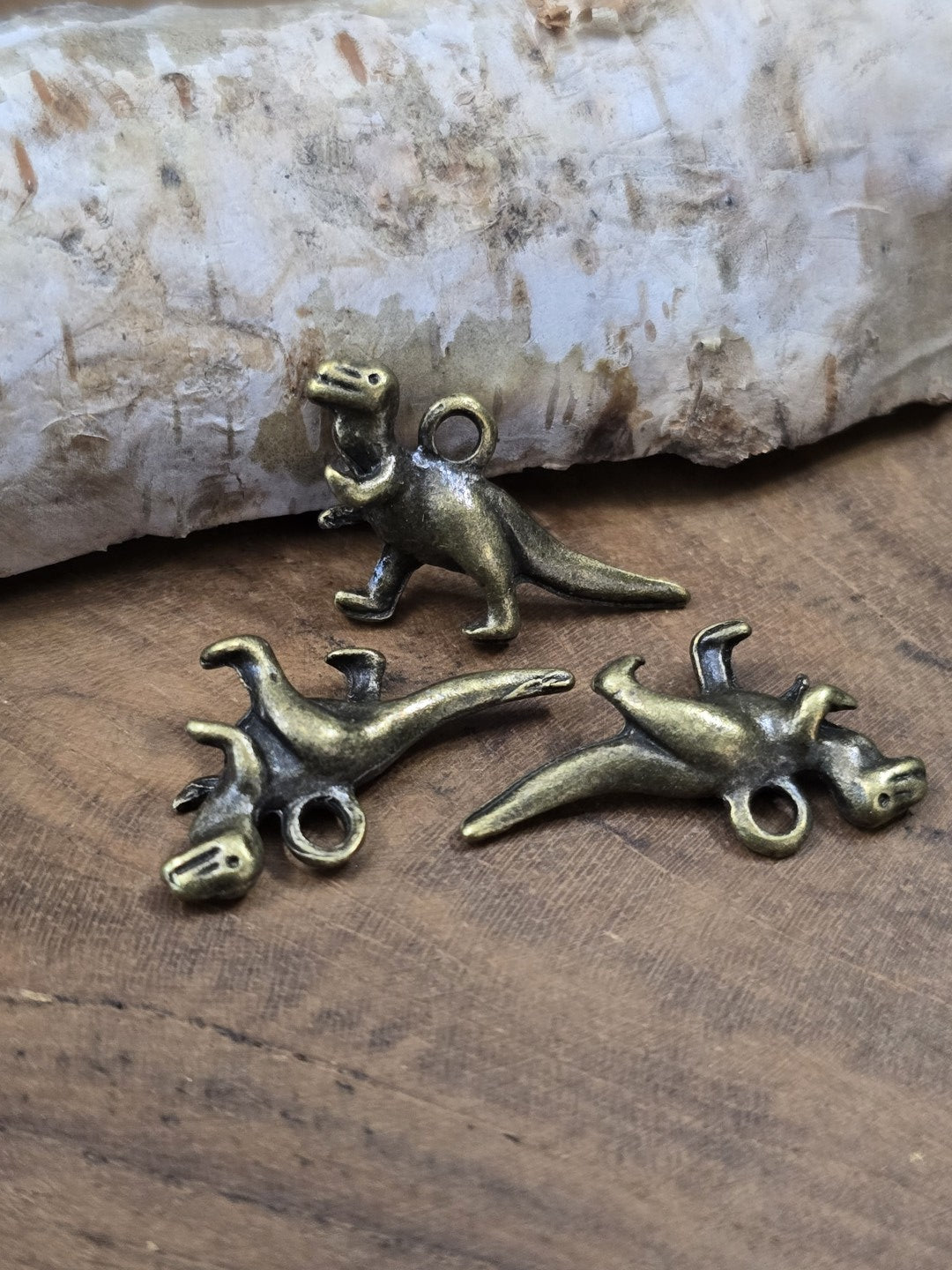 Drei detailreiche T-Rex Dinosaurier-Anhänger in Antik-Bronze (27 mm) dekorativ stehend und liegend auf Holzgrund vor Birkenrinde.