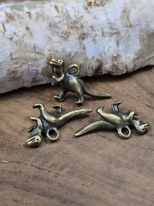 Drei detailreiche T-Rex Dinosaurier-Anhänger in Antik-Bronze (27 mm) dekorativ stehend und liegend auf Holzgrund vor Birkenrinde.