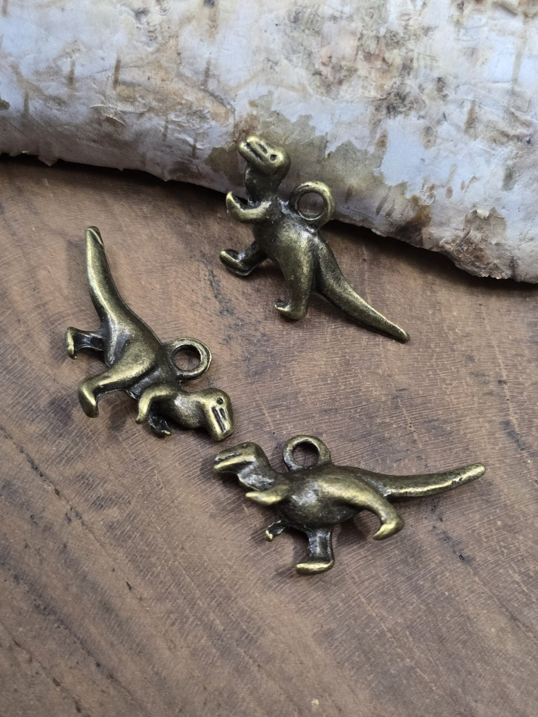 Drei urzeitliche T-Rex Charms in Antik-Bronze aus verschiedenen Winkeln auf einer Holzplatte zur Ansicht der plastischen Ausarbeitung.