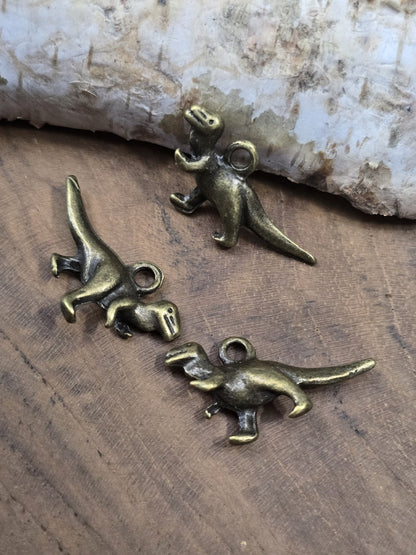 Drei urzeitliche T-Rex Charms in Antik-Bronze aus verschiedenen Winkeln auf einer Holzplatte zur Ansicht der plastischen Ausarbeitung.