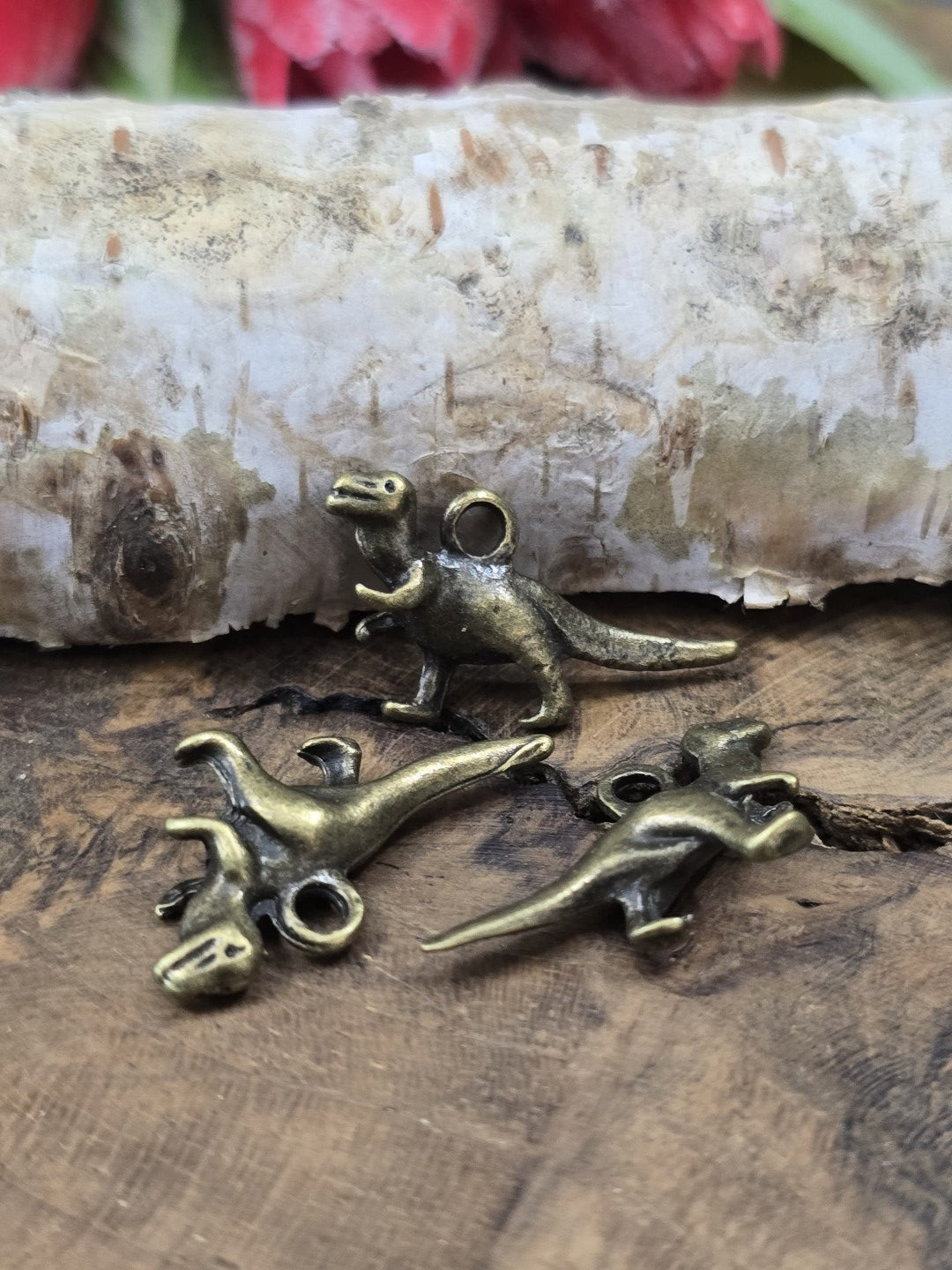 Drei T-Rex Dino-Schmuckanhänger (27 mm) aus Metall zum Basteln von Ketten und Schlüsselanhängern auf rustikalem Holzgrund.