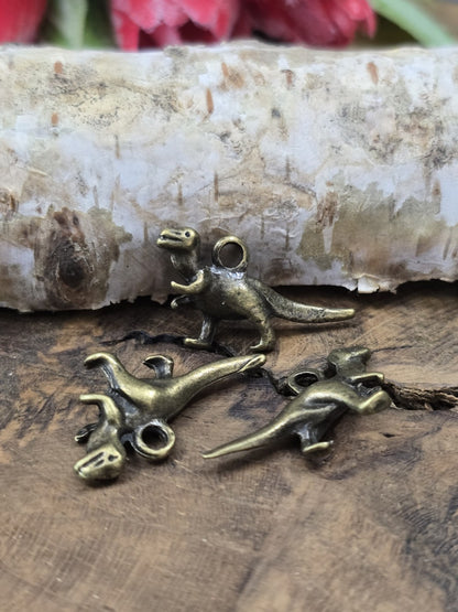 Drei T-Rex Dino-Schmuckanhänger (27 mm) aus Metall zum Basteln von Ketten und Schlüsselanhängern auf rustikalem Holzgrund.