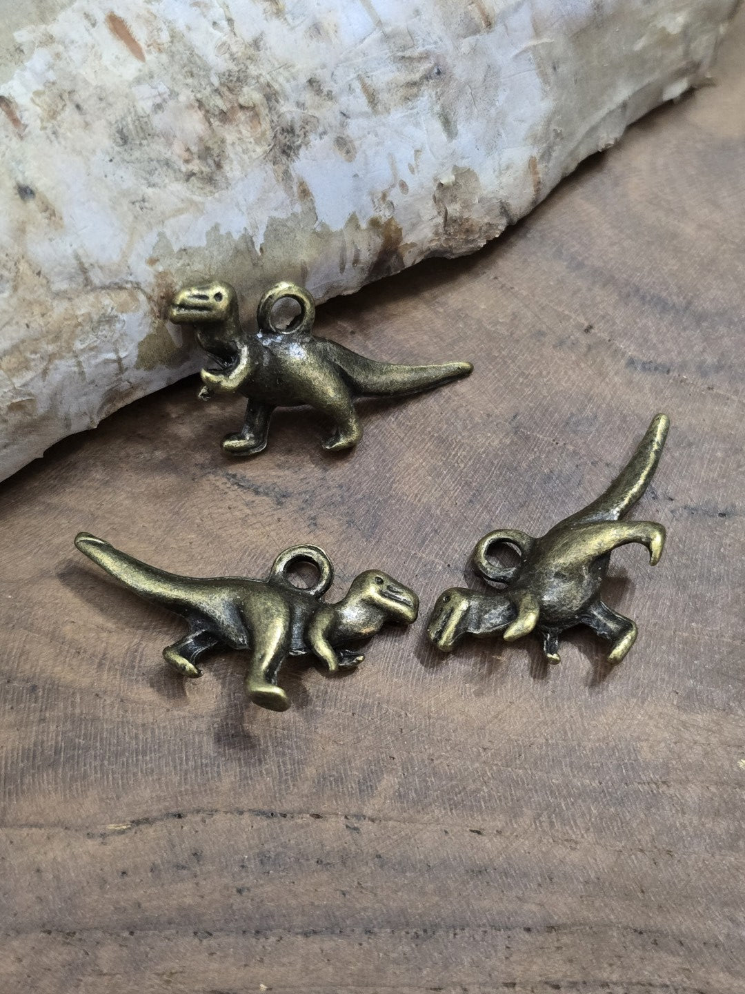 Drei antik-bronzefarbene Dinosaurier-Anhänger flach liegend auf Holz zur Veranschaulichung der Vorder- und Rückseite sowie der Aufhängeöse.