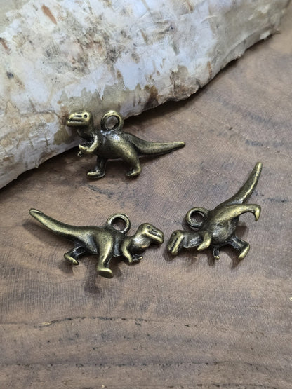 Drei antik-bronzefarbene Dinosaurier-Anhänger flach liegend auf Holz zur Veranschaulichung der Vorder- und Rückseite sowie der Aufhängeöse.