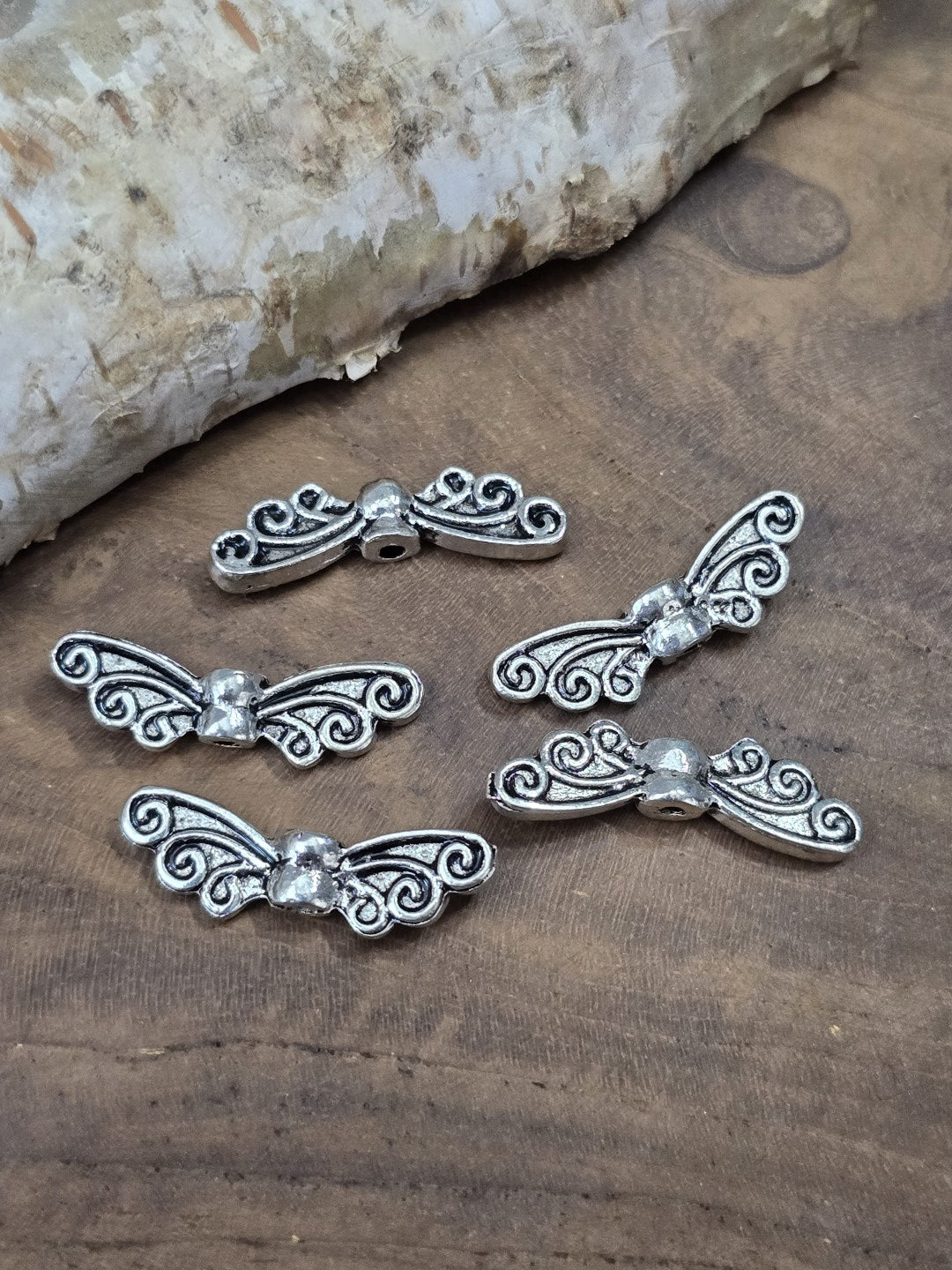 Fünf filigrane Engelsflügel-Perlen in Antik-Silber (22 mm) flach liegend auf Holzgrund vor Birkenrinde.