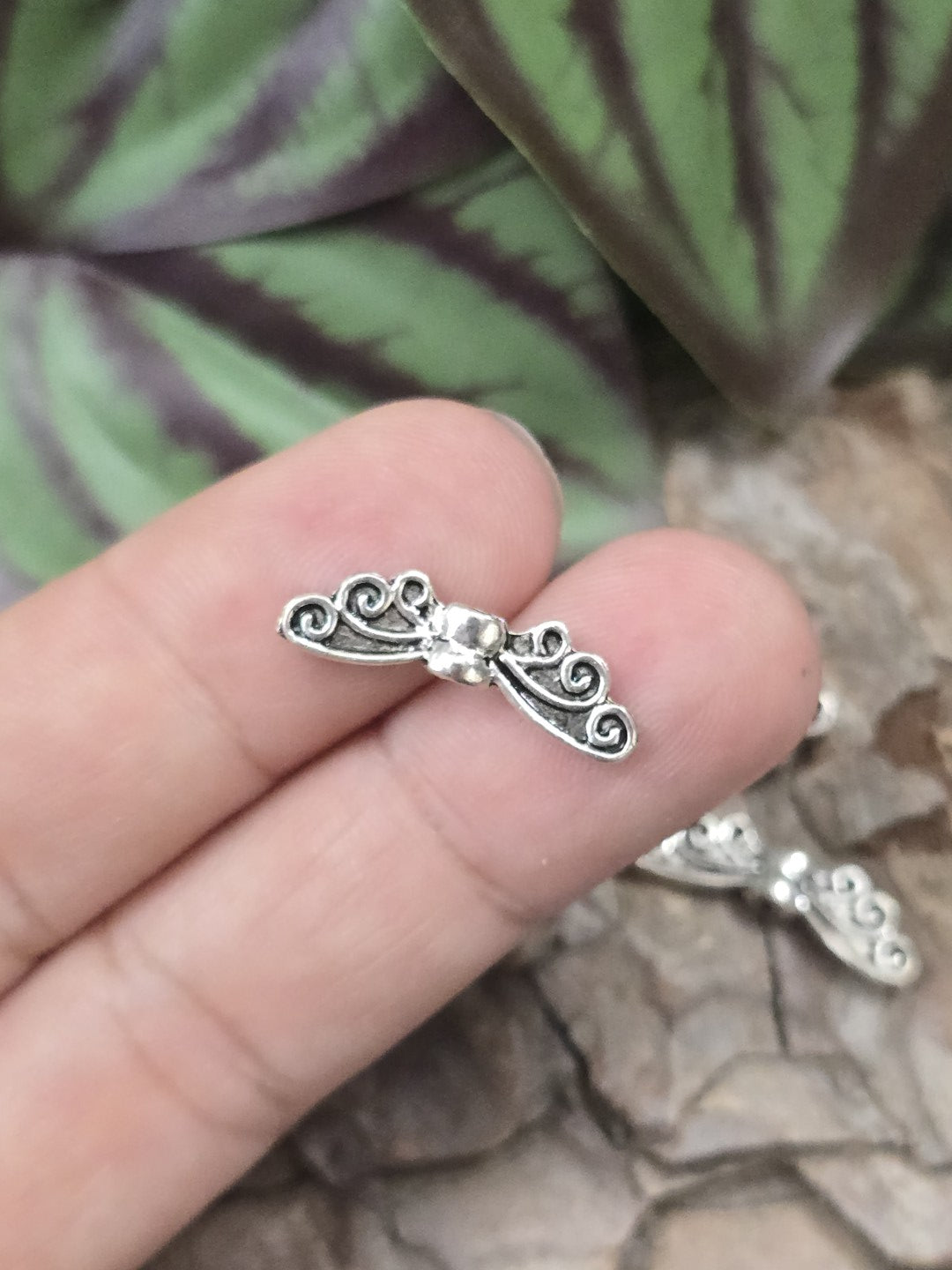 Eine kleine Engelsflügel-Zwischenperle in Antik-Silber auf einer Fingerkuppe präsentiert als Größenvergleich.
