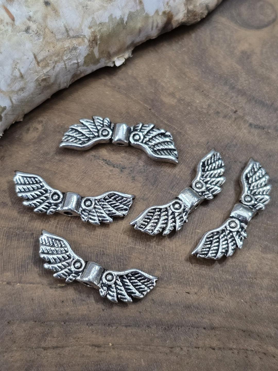 Fünf filigrane Engelsflügel-Perlen mit feinem Federmuster in Antik-Silber (22 mm) flach liegend auf Holzgrund vor Birkenrinde.