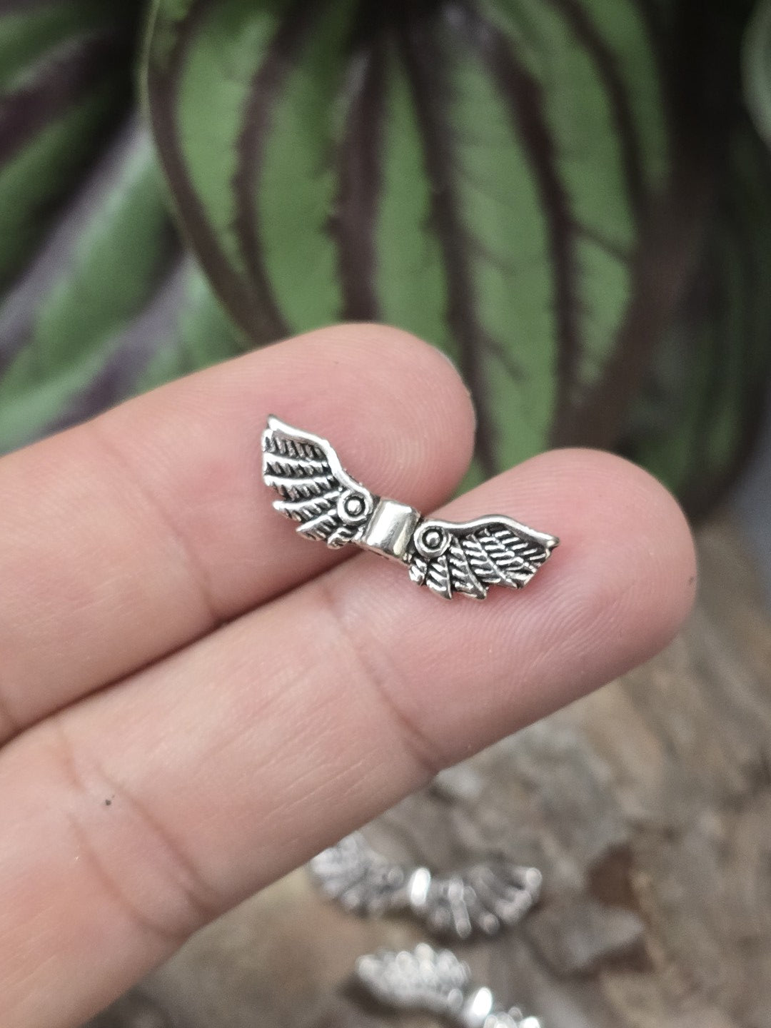 Eine kleine Feder-Engelsflügel-Zwischenperle in Antik-Silber auf einer Fingerkuppe präsentiert als Größenvergleich.