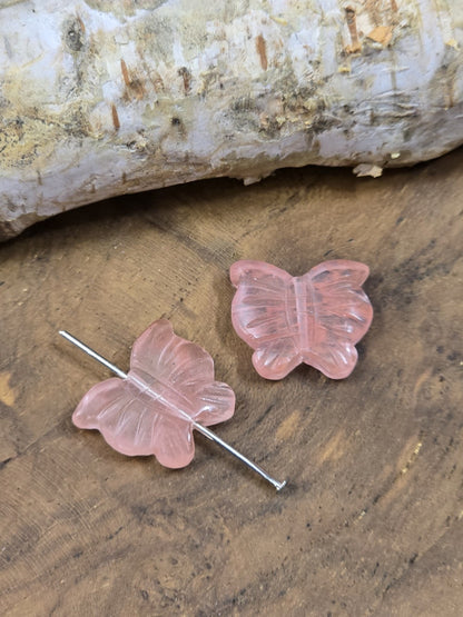 Perle aus Erdbeerquarz mit Nietstift zur Veranschaulichung des Verlaufs des Fädellochs, daneben liegt eine weitere Perle in Schmetterlingsform aus Erdbeerquarz.
