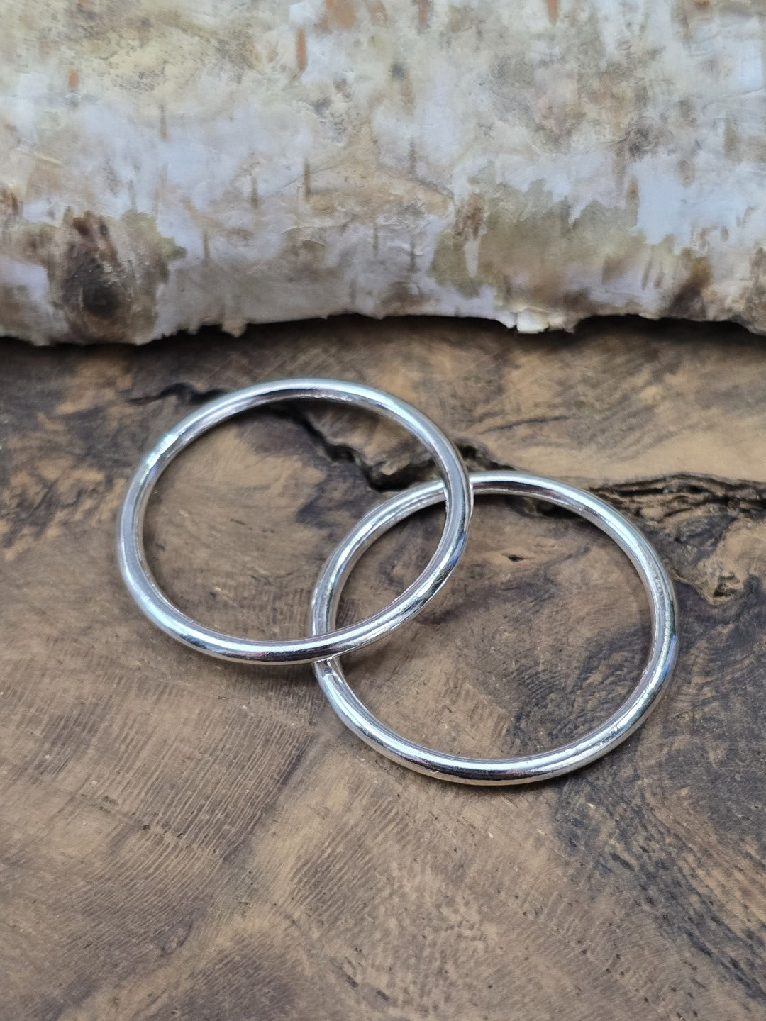 Zwei glänzende, geschlossene Metallringe in Silber (24 mm) dekorativ ineinandergelegt auf einer rustikalen Holzoberfläche vor Birkenrinde.