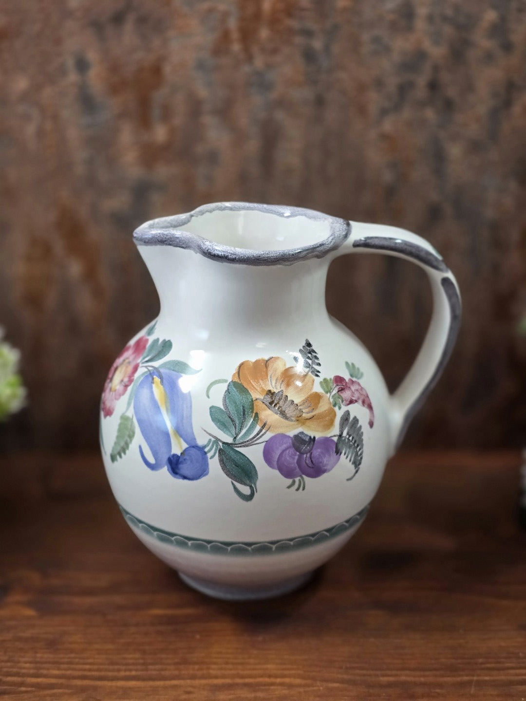 Vintage Keramikkrug mit farbigem Blumendekor, großem Henkel und geschwungenem Ausguss auf dunkler Holzfläche in Frontalansicht.