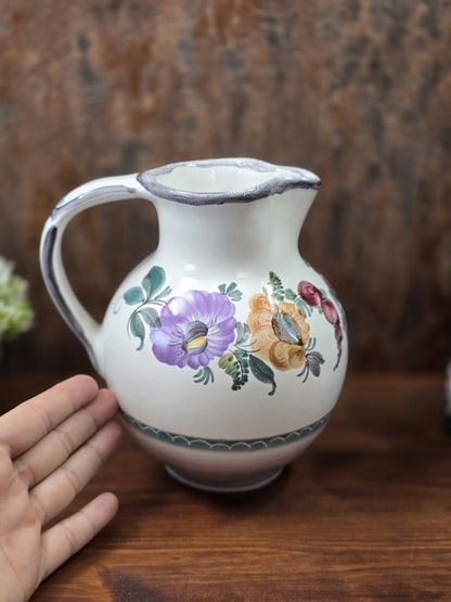Vintage Keramikkrug mit violetten und ockerfarbenen Blüten, grünem Blattdekor und grauem Rand, neben einer Hand als Größenvergleich.