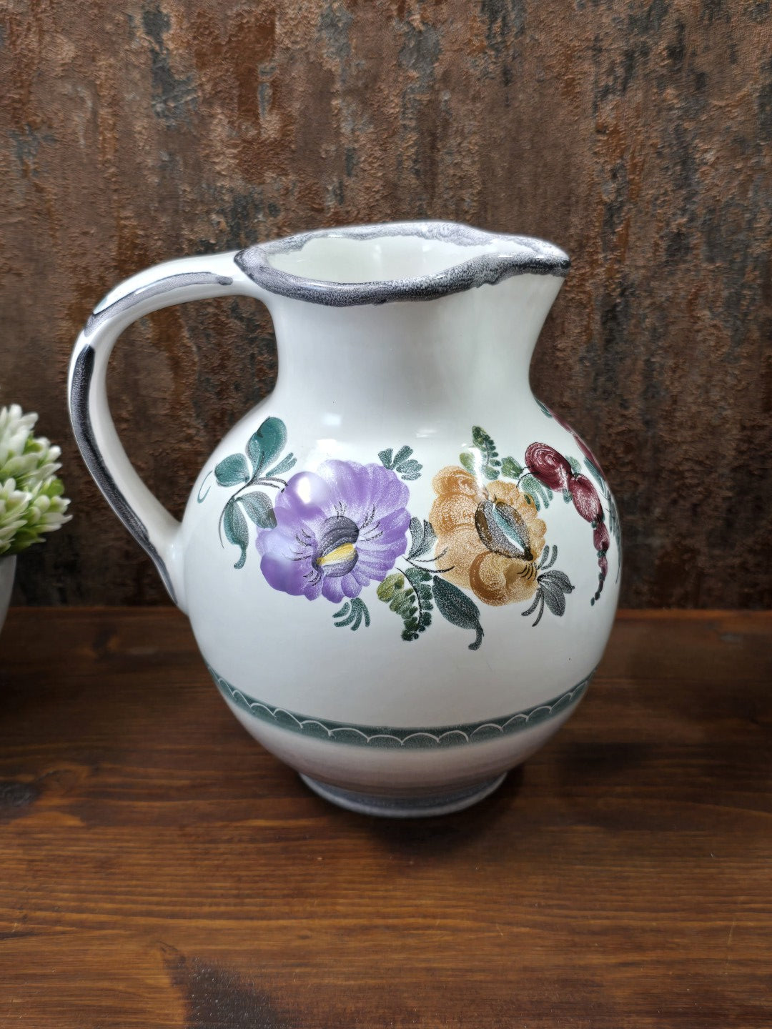 Vintage Keramikkrug mit großem Henkel und floralem Dekor in Violett, Ocker, Rot und Grün auf dunklem Holz vor braunem Hintergrund.