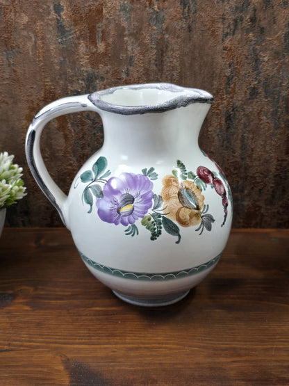 Vintage Keramikkrug mit großem Henkel und floralem Dekor in Violett, Ocker, Rot und Grün auf dunklem Holz vor braunem Hintergrund.