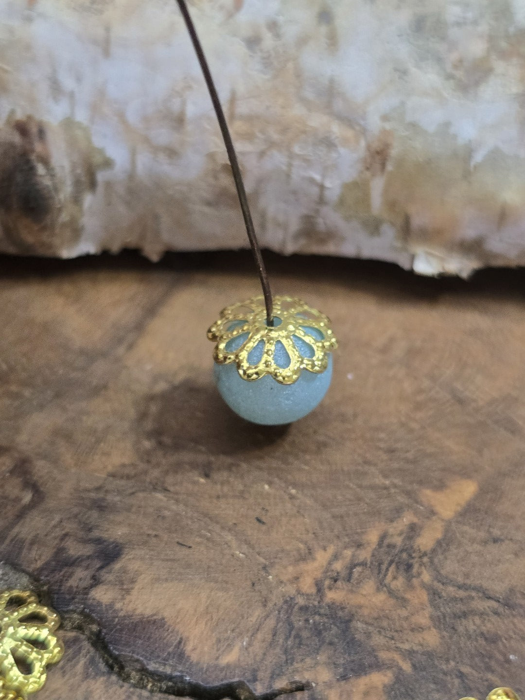 Goldene blütenförmige Perlenkappe auf einer hellblauen Glaskugel aufgefädelt auf einem Nietstift.