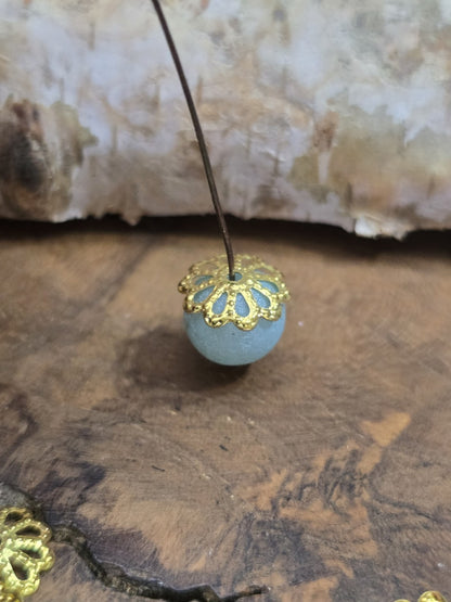 Goldene blütenförmige Perlenkappe auf einer hellblauen Glaskugel aufgefädelt auf einem Nietstift.