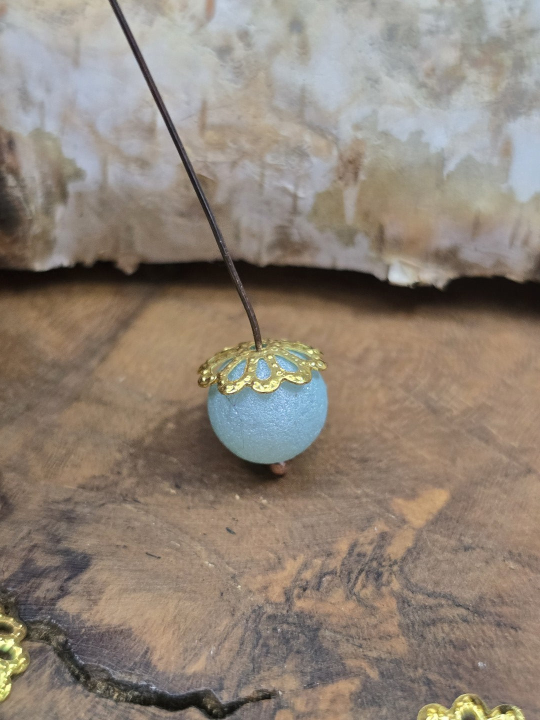 Seitenansicht einer filigranen Perlenkappe in Gold als Verzierung auf einer hellblauen Schmuckperle.