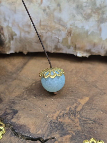 Seitenansicht einer filigranen Perlenkappe in Gold als Verzierung auf einer hellblauen Schmuckperle.